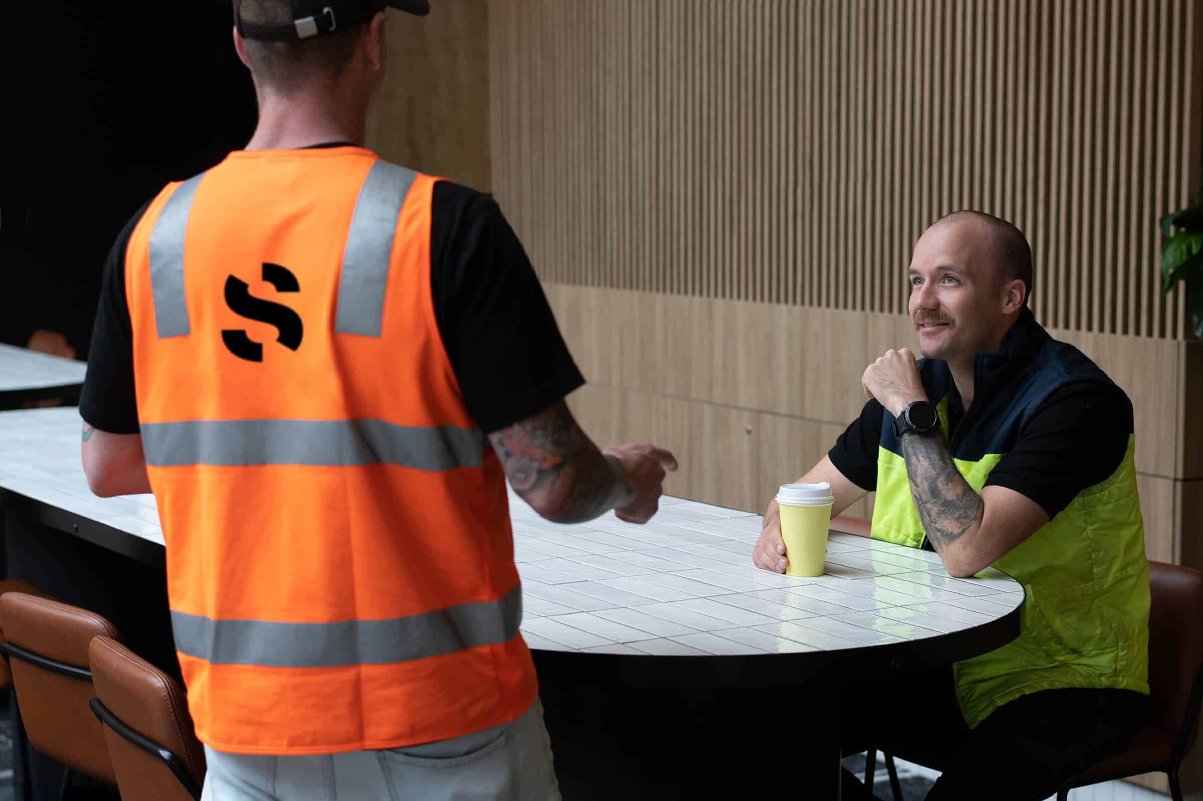 spaceful-team-construction Two men in construction uniforms chatting over a coffee.
