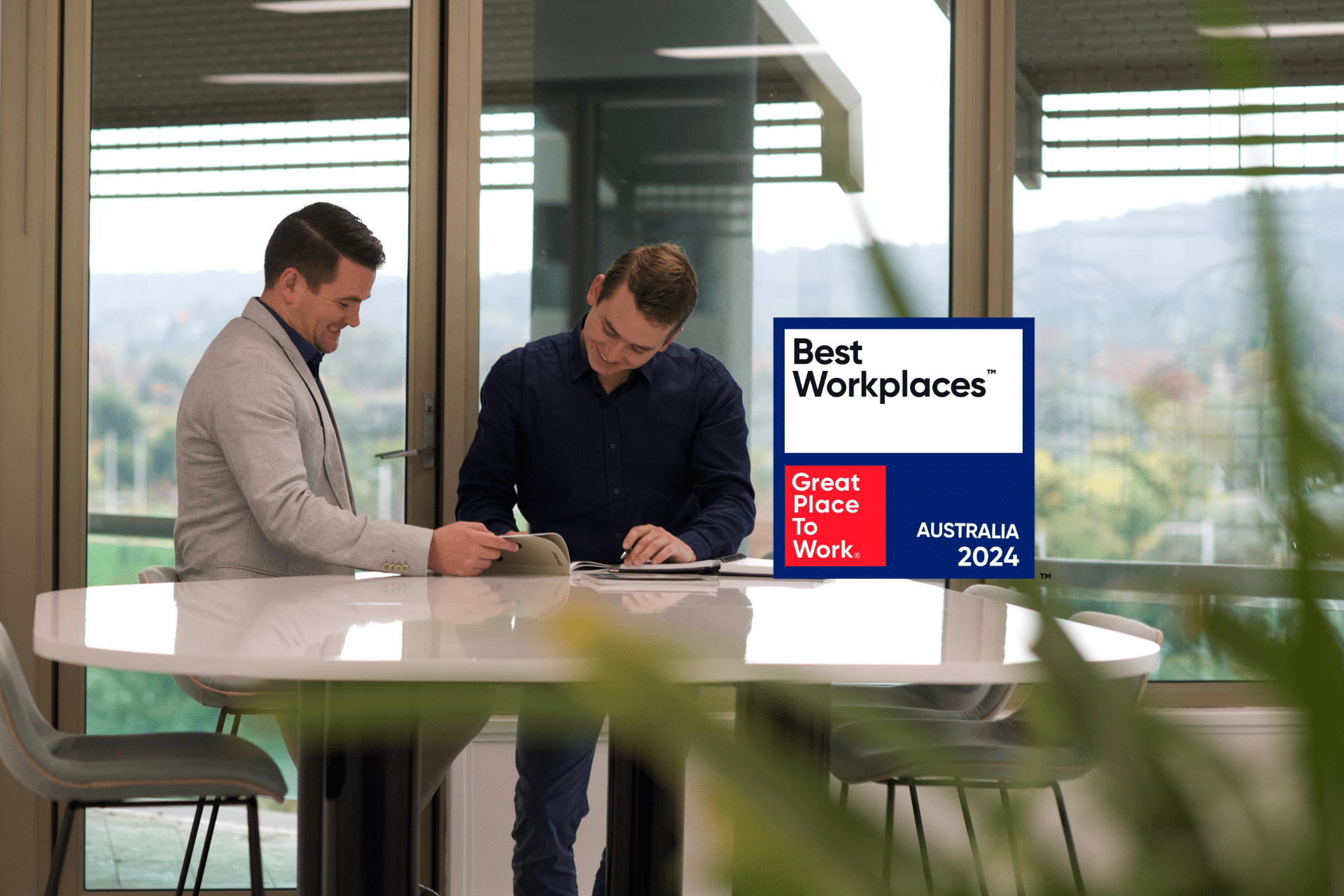 Two people smiling, looking over a booklet in an open plan office with large windows and natural light. Best Workplaces award in the foreground.